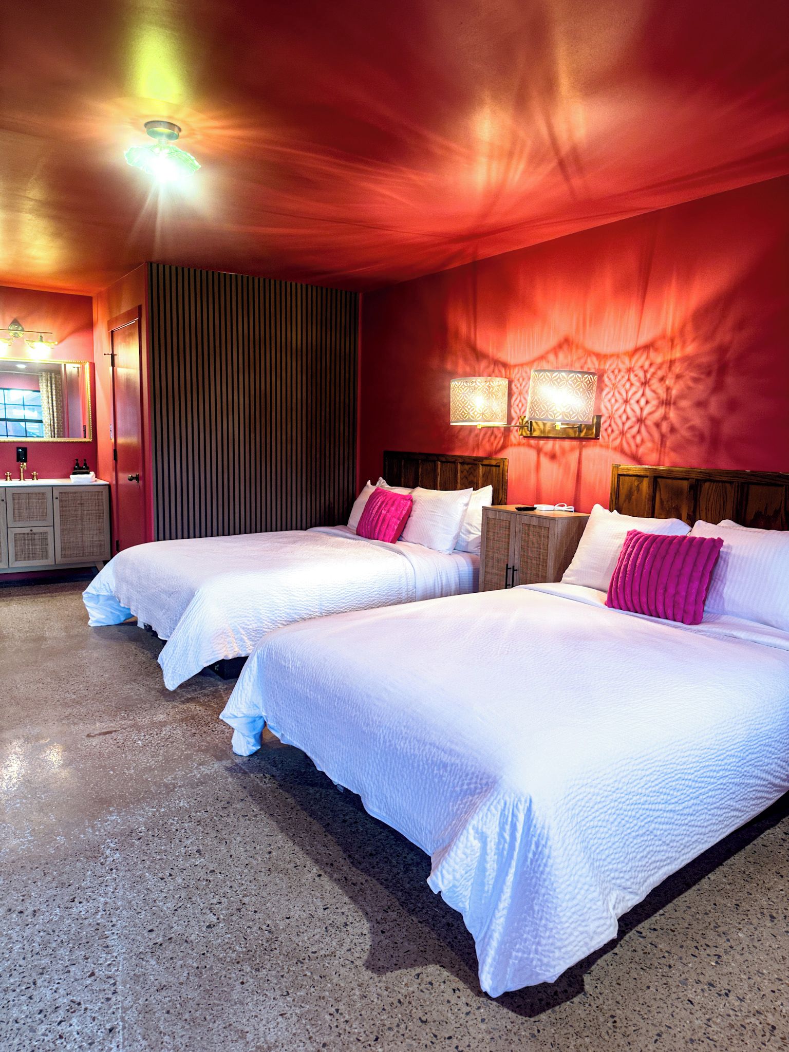 A hotel room with two beds, a vanity with a mirror, and a red accent wall.