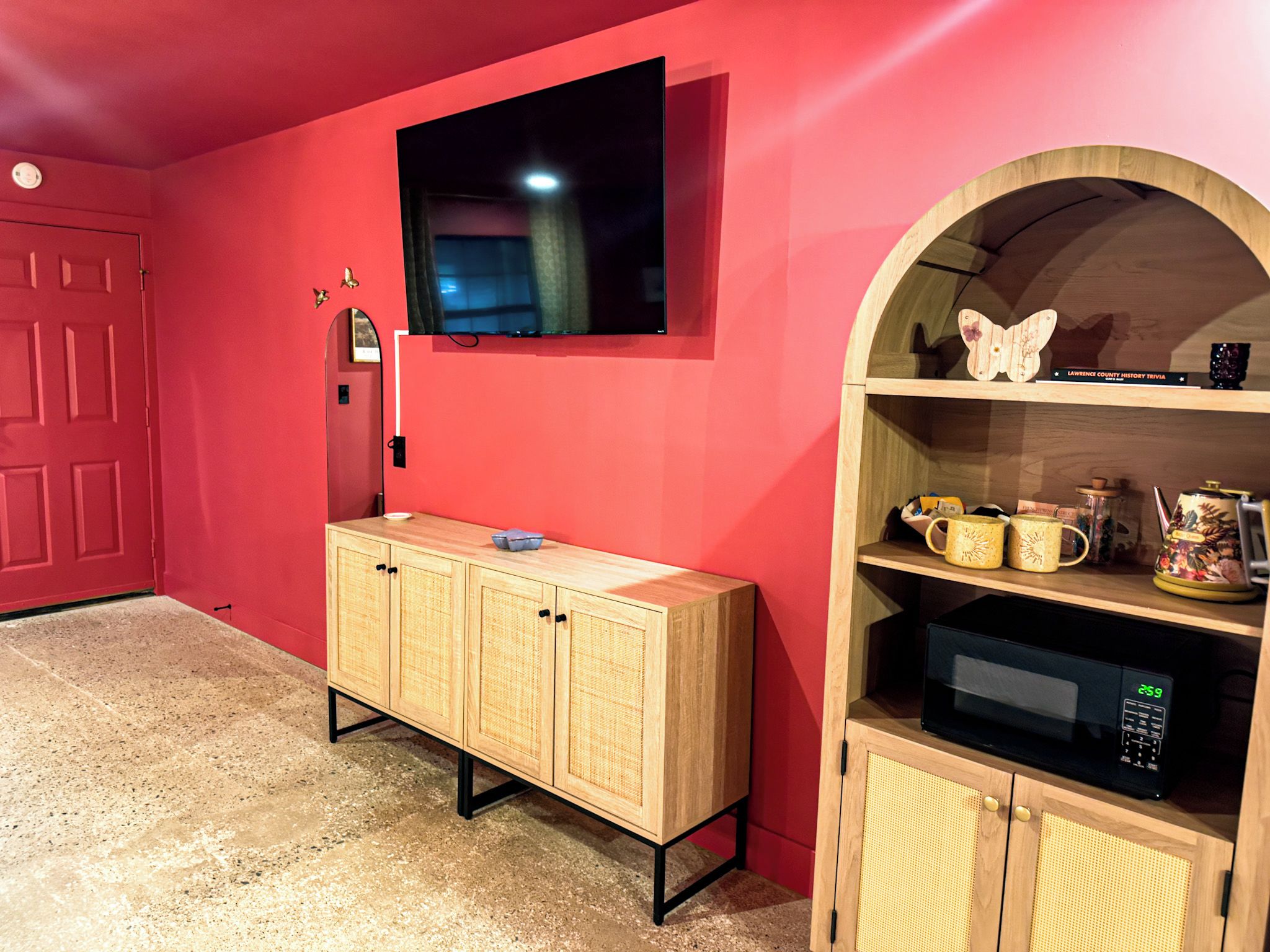 Una habitación con una pared roja, un armario de madera con un televisor de pantalla plana montado encima, y un estante de madera con objetos decorativos.