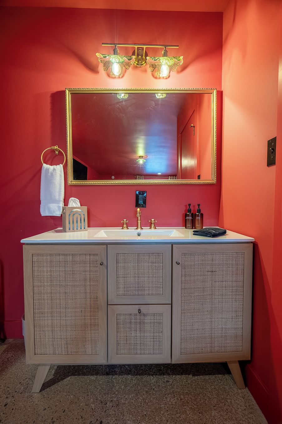 A bathroom with a red wall, a large mirror with a gold frame, a white sink, and a wooden cabinet with mesh doors.