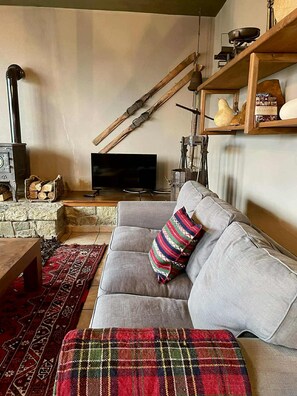 A cozy living room with a stone fireplace, a television, a sofa with a plaid throw, and a wooden shelf with decorative items.