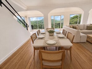 Uma sala de jantar com mesa posta para quatro pessoas, rodeada por cadeiras de vime, com uma varanda e um sofá ao fundo.