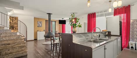 A modern kitchen with a central island, a dining area with chairs, a staircase, and a living room with a fireplace.