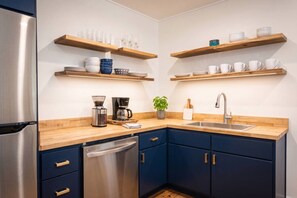 A modern kitchen with a stainless steel refrigerator, wooden shelves with dishes, a coffee maker, and a sink. - Two living rooms, cowboy pool and fire pit (Austin)