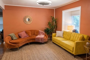 A living room with a curved orange sofa, a yellow sofa, a round mirror, and a large window with blinds. - Two living rooms, cowboy pool and fire pit (Austin)