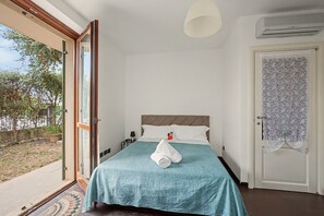 A hotel room with a bed, a nightstand, a lamp, a window with a view of greenery, and an air conditioning unit.