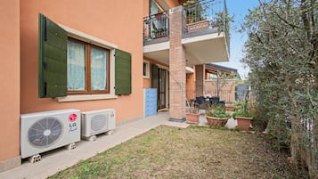 Een woongebouw met een balkon over twee verdiepingen, een klein tuintje met plantenpotten en twee airconditioners op de begane grond.