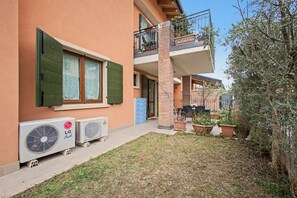 A residential building with a two-story balcony, a small garden area with potted plants, and two air conditioning units on the ground floor.