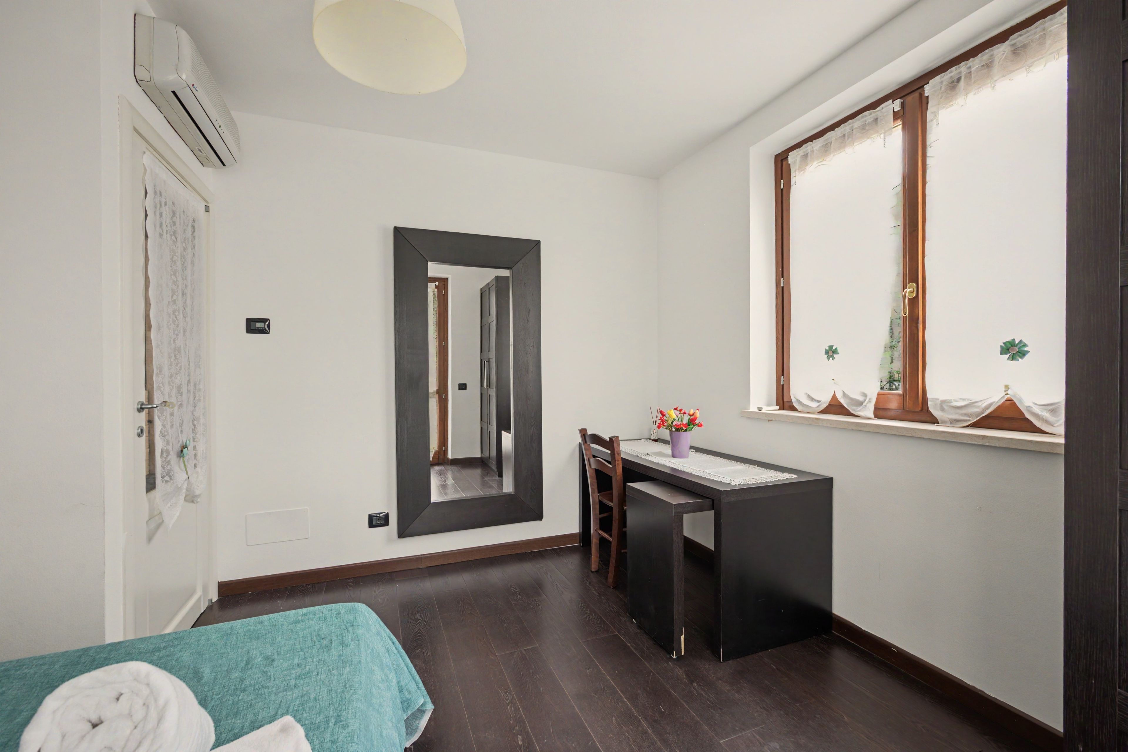 Ein Zimmer mit einem Bett, einem Schreibtisch, einem Stuhl, einem Vorhangfenster und einem Spiegel an der Wand.