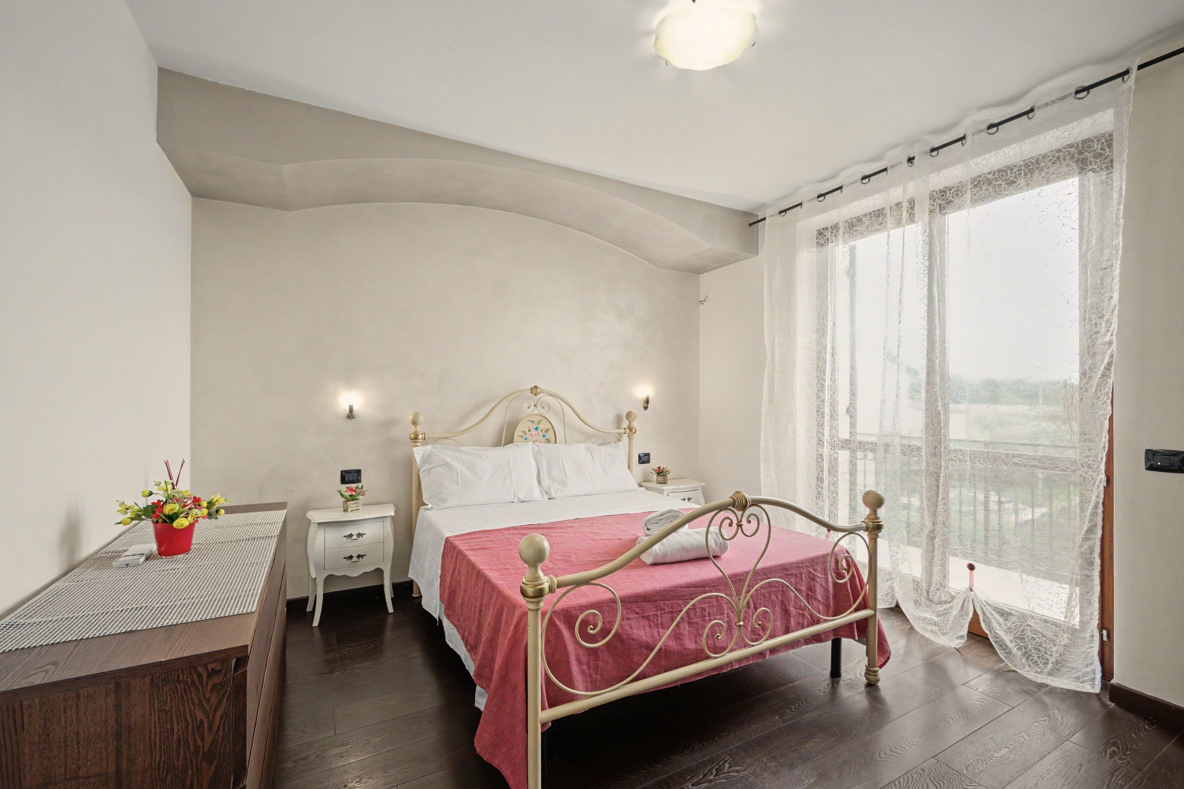 Ein Schlafzimmer mit Bett, Nachttischen, einem Fenster mit Spitzenvorhängen und Blick auf die Stadt.