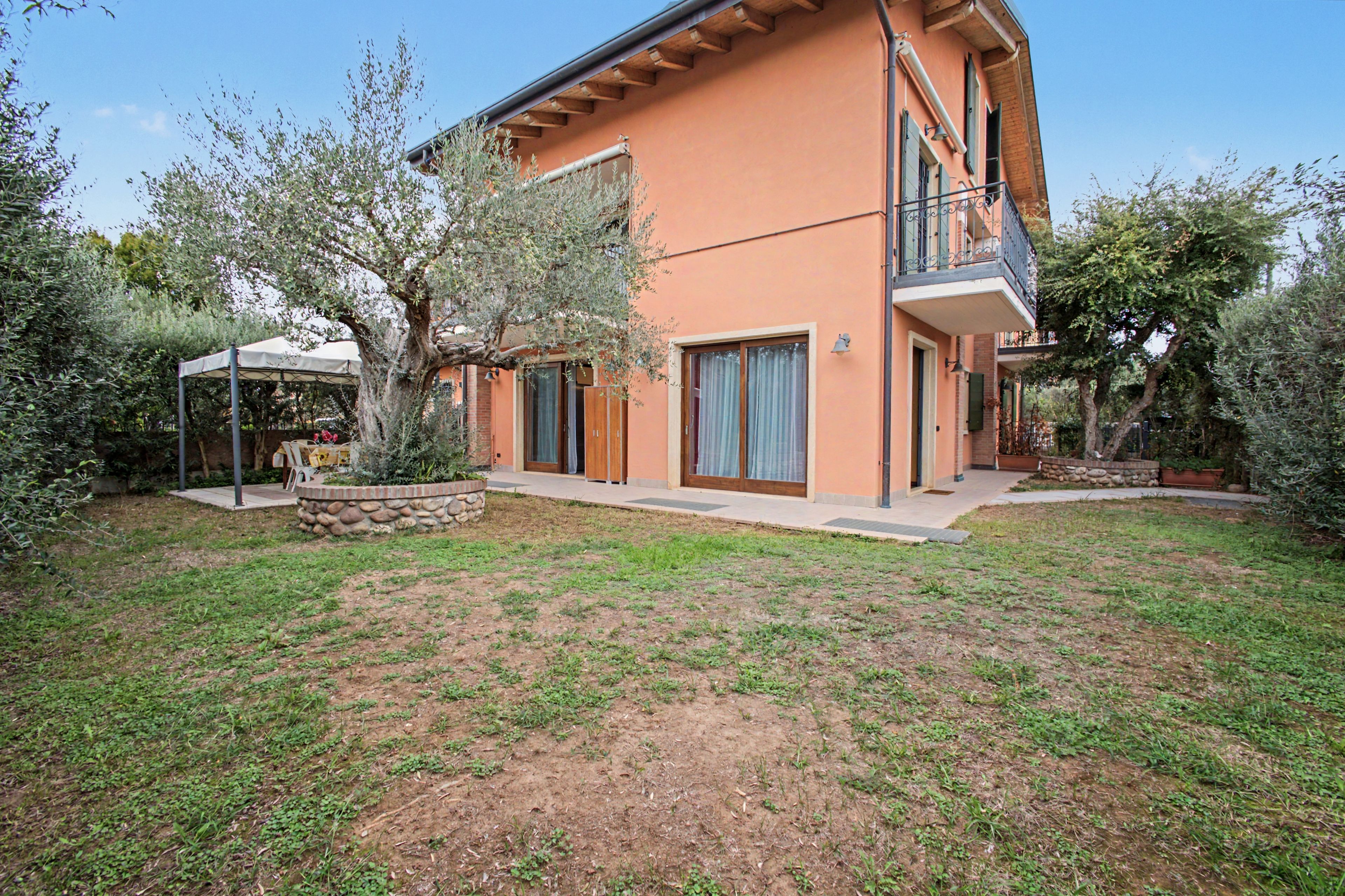 Una casa a due piani con fondamenta in pietra, un balcone e un giardino alberato con un piccolo stagno.
