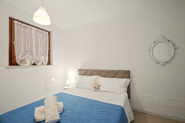 A bedroom with a bed, a window with curtains, a bedside lamp, a wall-mounted mirror, and a towel on the bed.