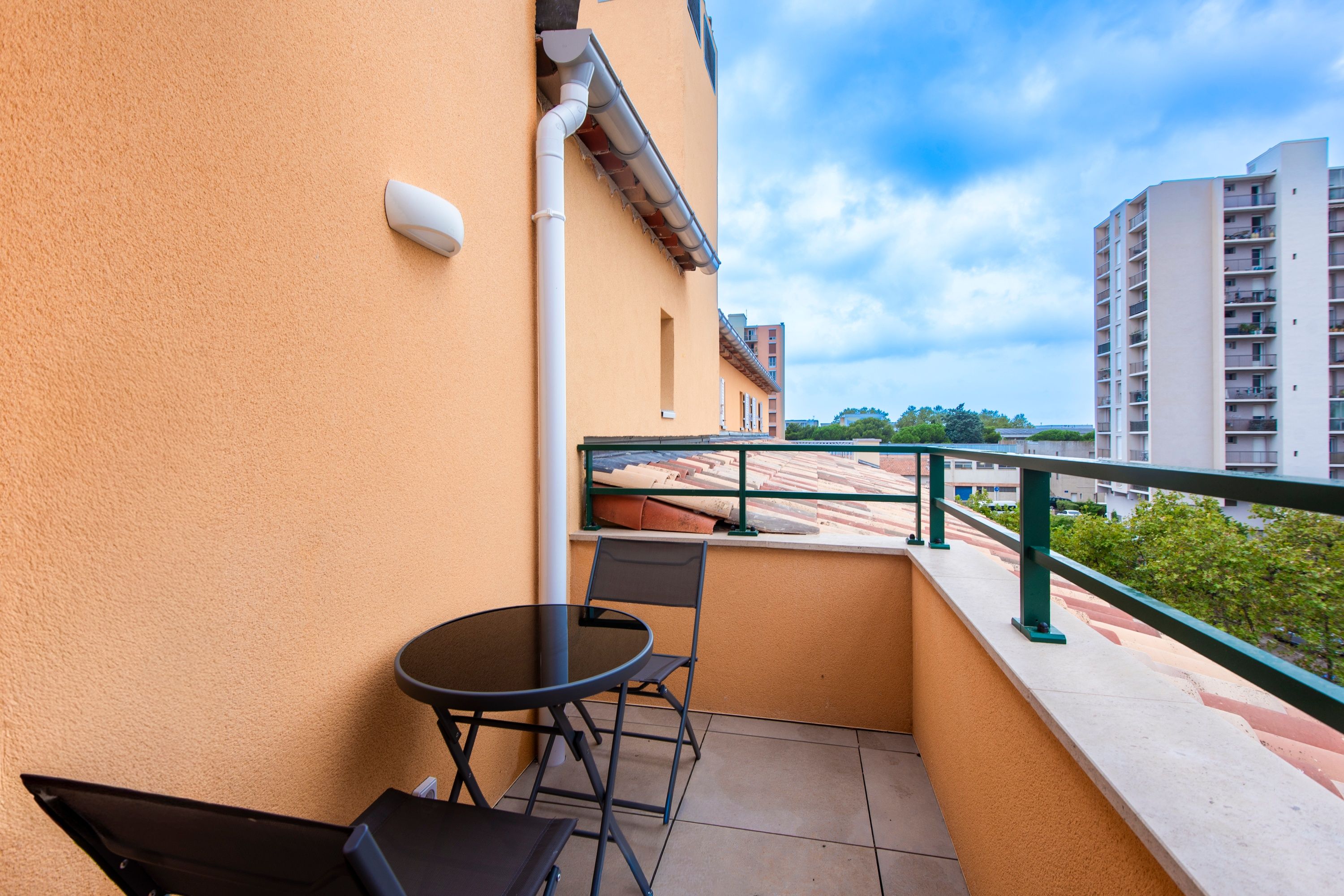 Un balcon avec une table et des chaises, une balustrade et une vue sur des immeubles et des arbres.