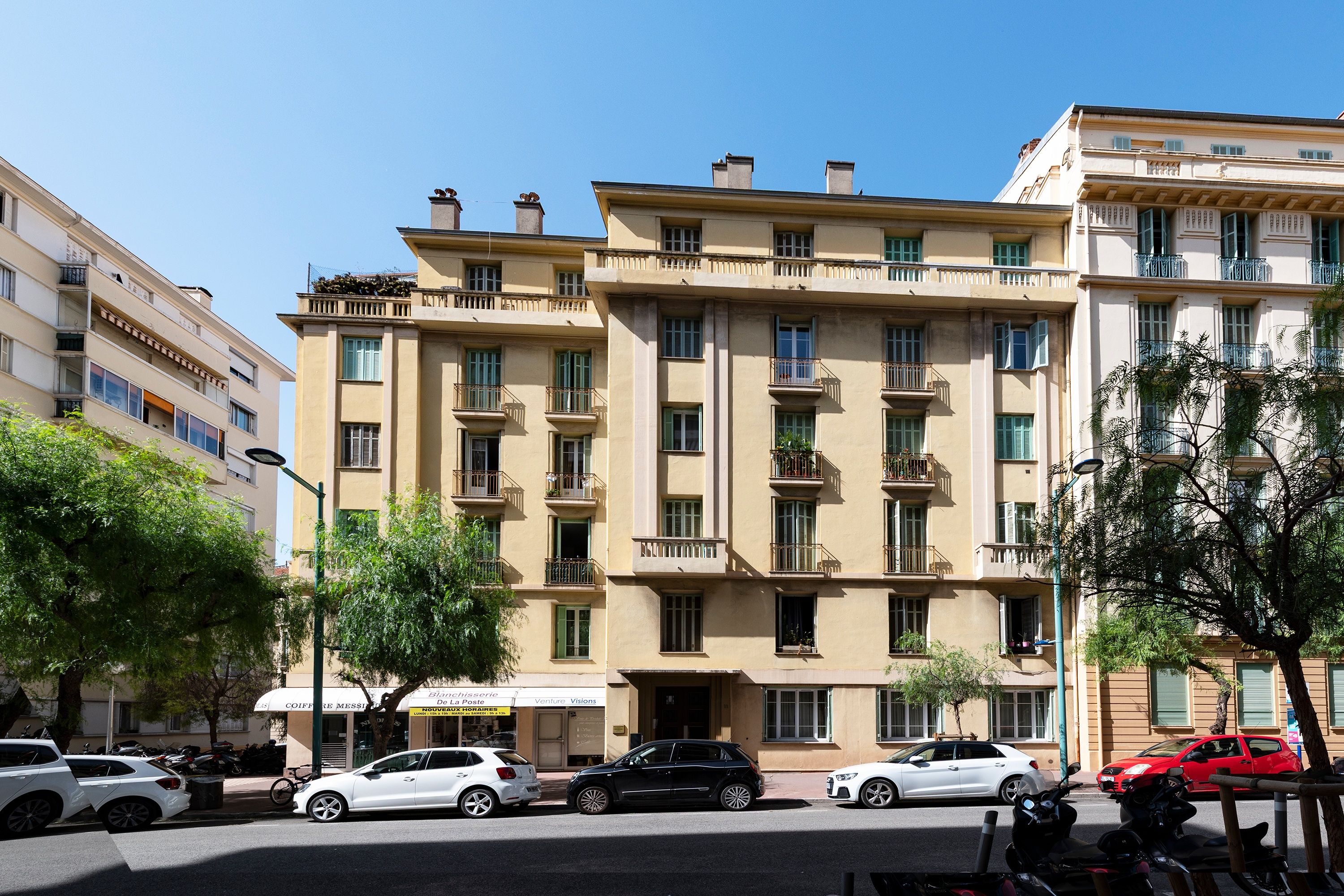 A multi-story building with balconies, parked cars, and a clear sky.