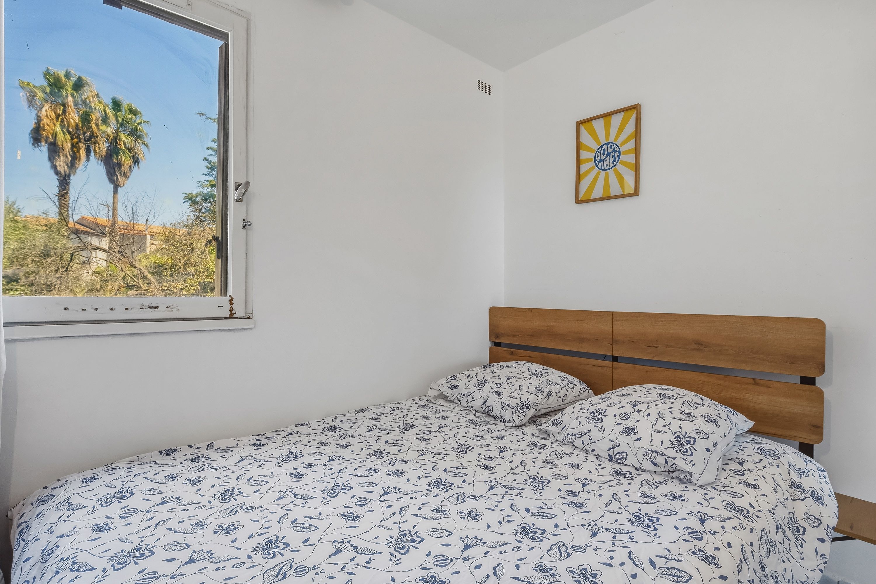 A bedroom with a single bed, a window with a view of palm trees, and a wall-mounted picture frame.