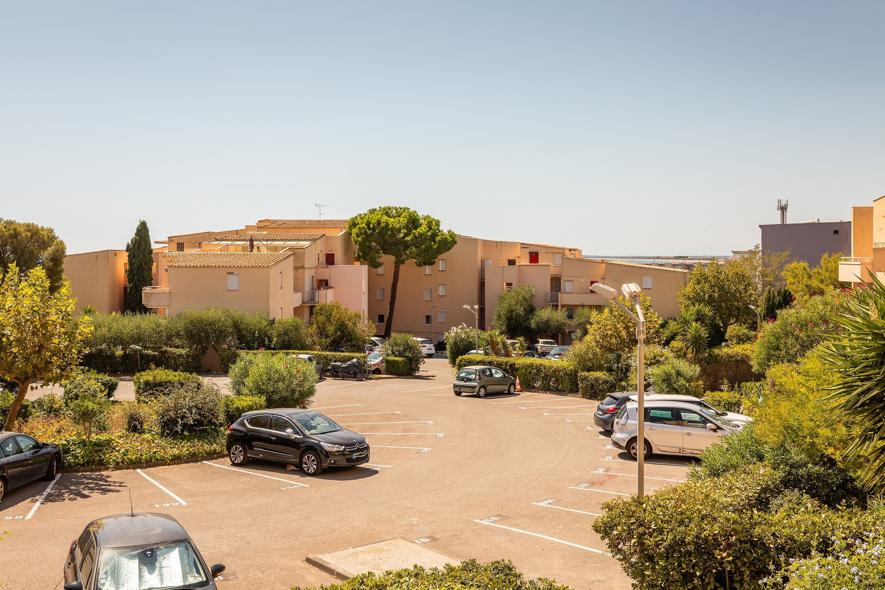 Un estacionamiento con varios autos, rodeado de vegetación y edificios de estilo mediterráneo.
