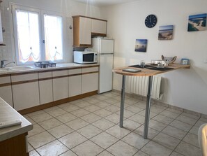 A kitchen with a sink, microwave, and cabinets. There is a table with a chair and a window with curtains. - L’Escale du Teich - à 2 km de la gare (Le Teich)