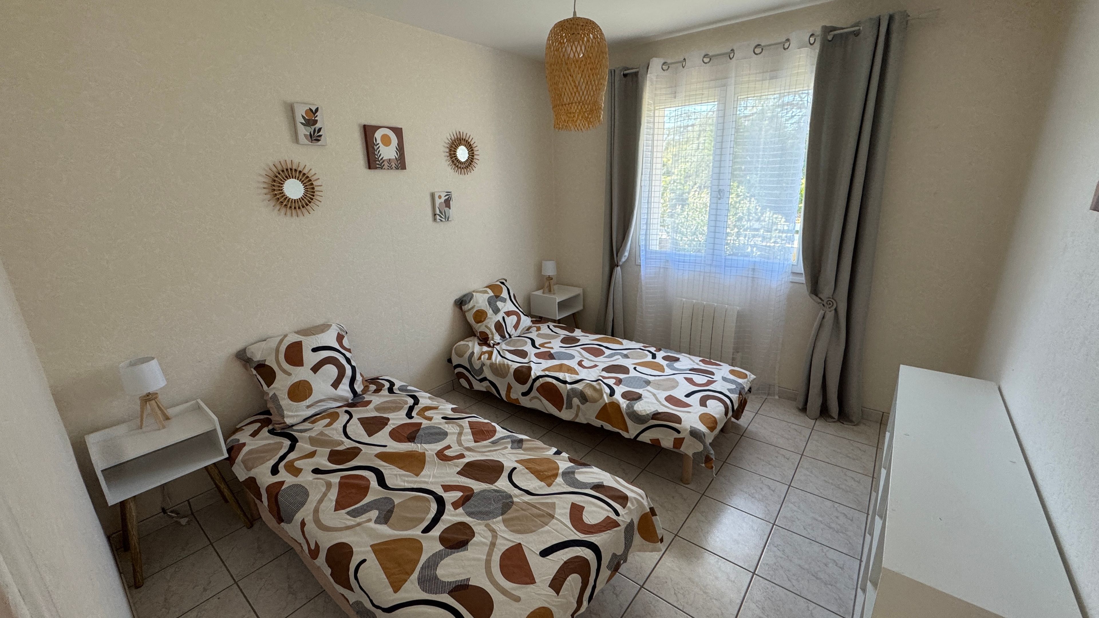 Une chambre avec deux lits, une table de chevet avec une lampe, un luminaire suspendu et une fenêtre avec des rideaux.
