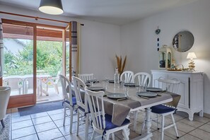 A dining area with a table set for four, white chairs, a white cabinet with a mirror, and a view of greenery outside. - L’Escale du Teich - à 2 km de la gare (Le Teich)