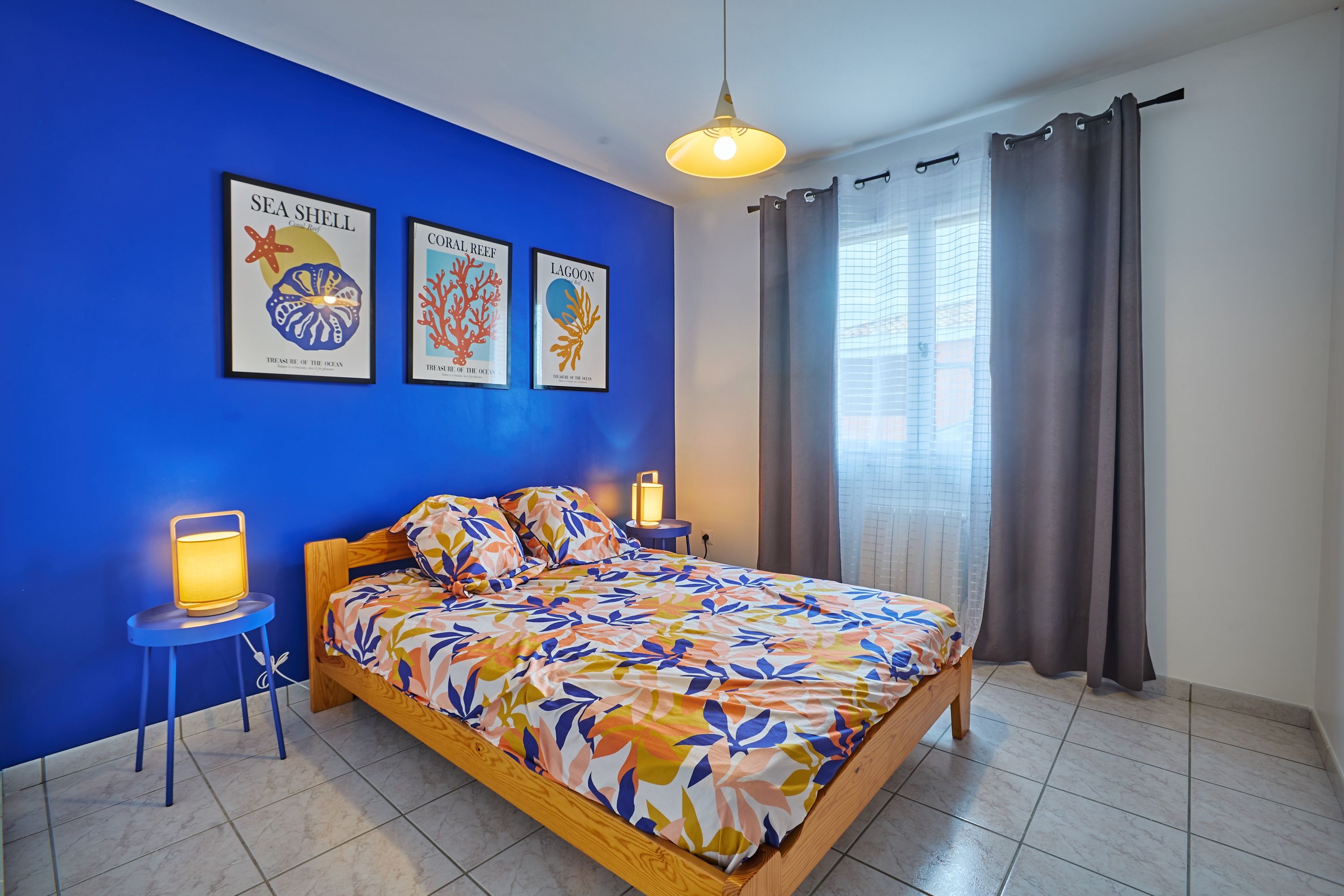 A bedroom with a bed, two bedside tables, a hanging light, and three framed posters on the wall.