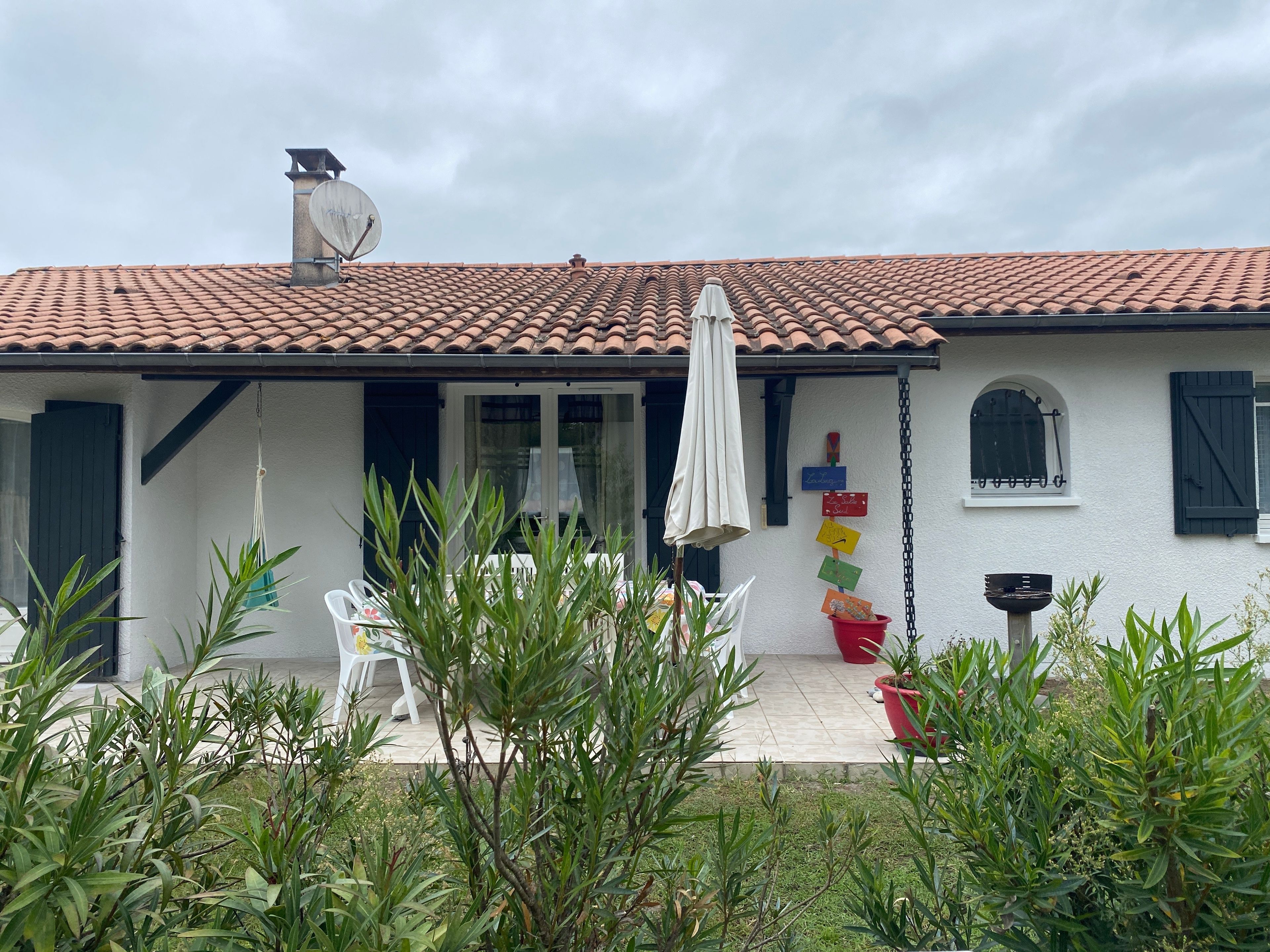 A house with a tiled roof, a satellite dish, a covered patio with chairs, and a small garden area.