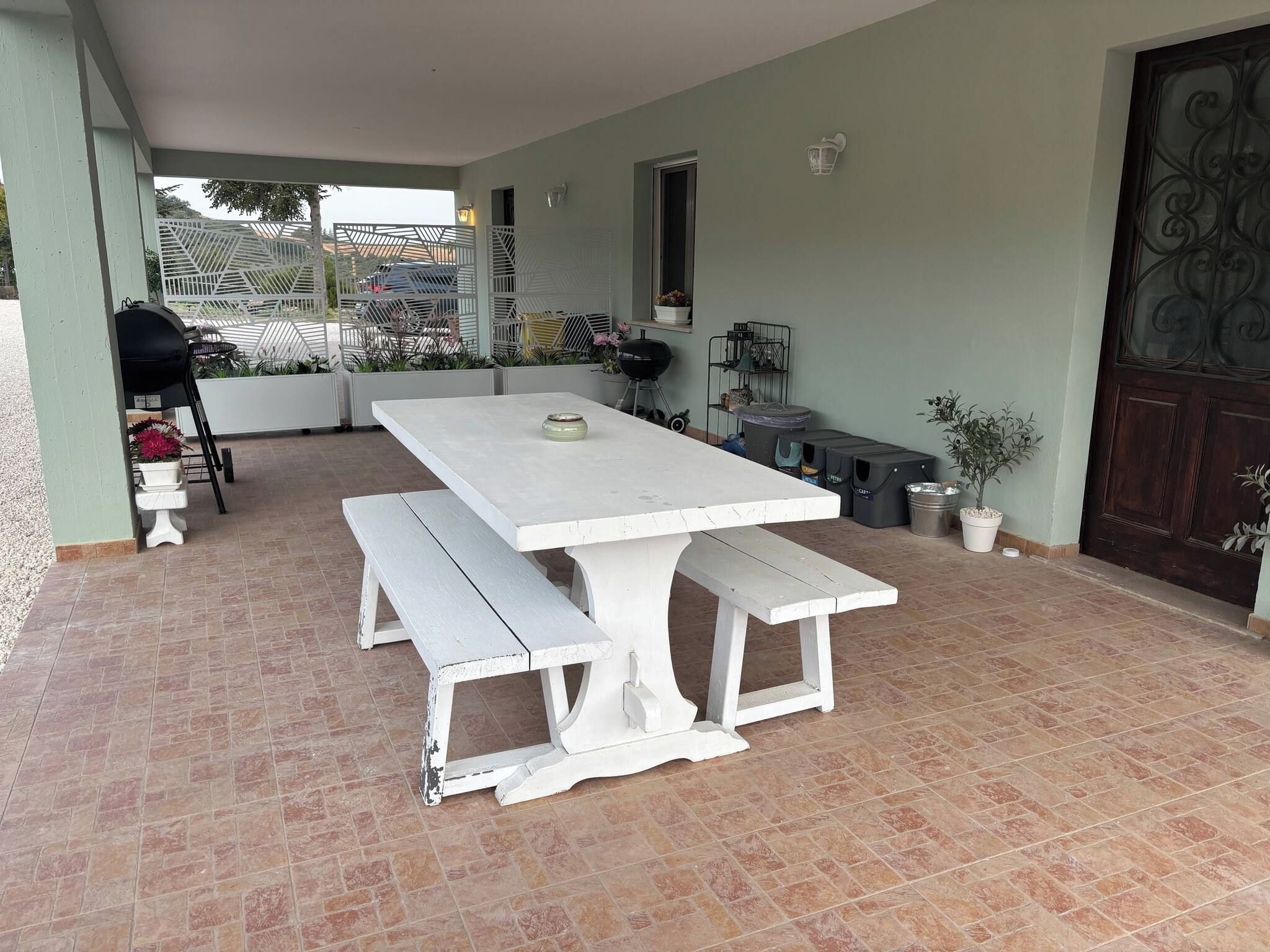 Um pátio com mesa e banco brancos, churrasqueira e plantas em vasos.
