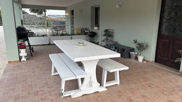 Um pátio com mesa e banco brancos, churrasqueira e plantas em vasos.