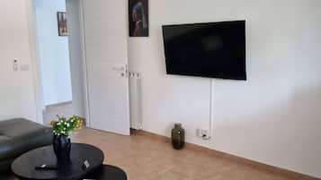 Un salon doté d’un ventilateur de plafond, d’un téléviseur à écran plat fixé au mur, d’une table basse ronde ornée d’un vase de fleurs et d’un canapé gris.