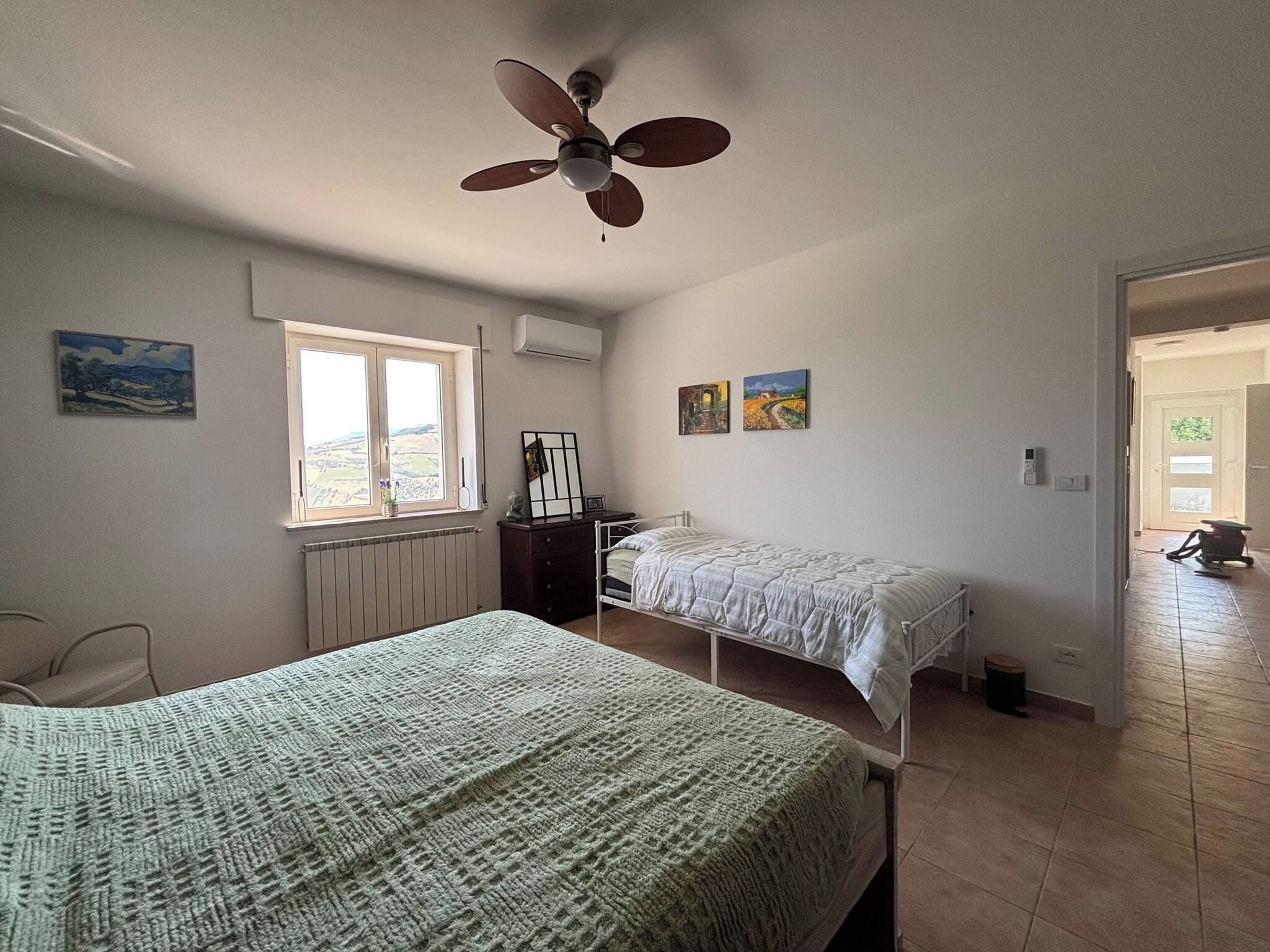 A bedroom with two beds, a ceiling fan, a window with a view, and artwork on the walls.