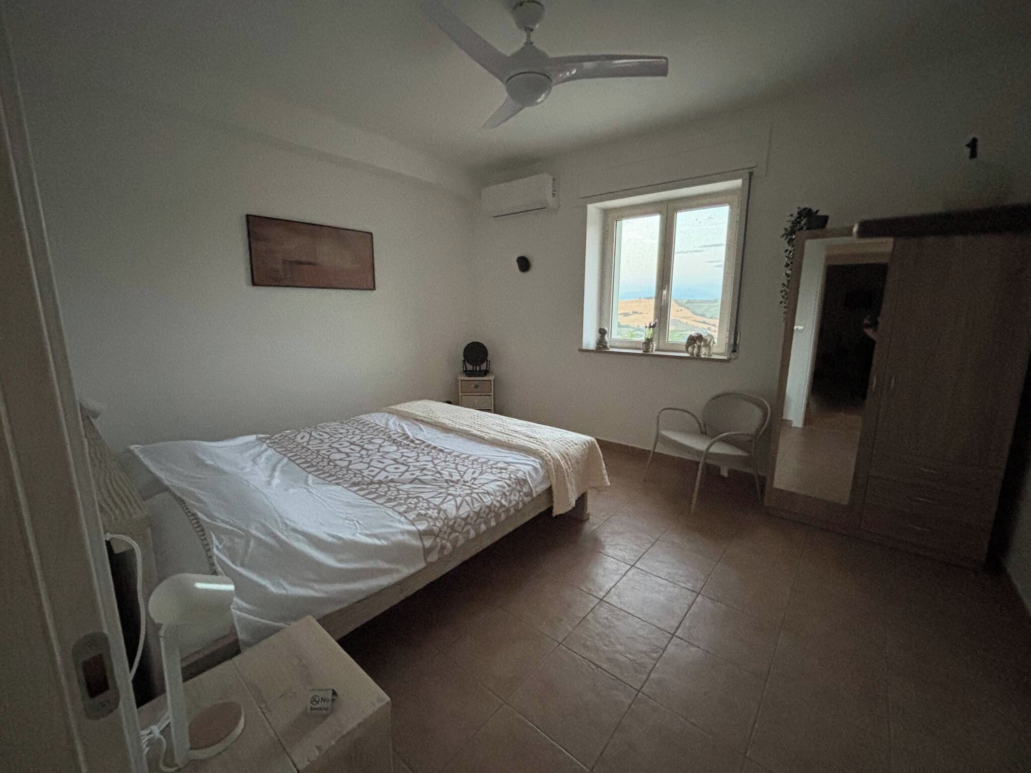 Quarto com cama, cadeira, escrivaninha, janela com vista e ventilador de teto.