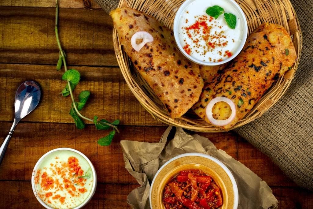 A wooden table with a basket containing two pieces of flatbread, a bowl of white sauce with red seasoning, and a bowl of red sauce.