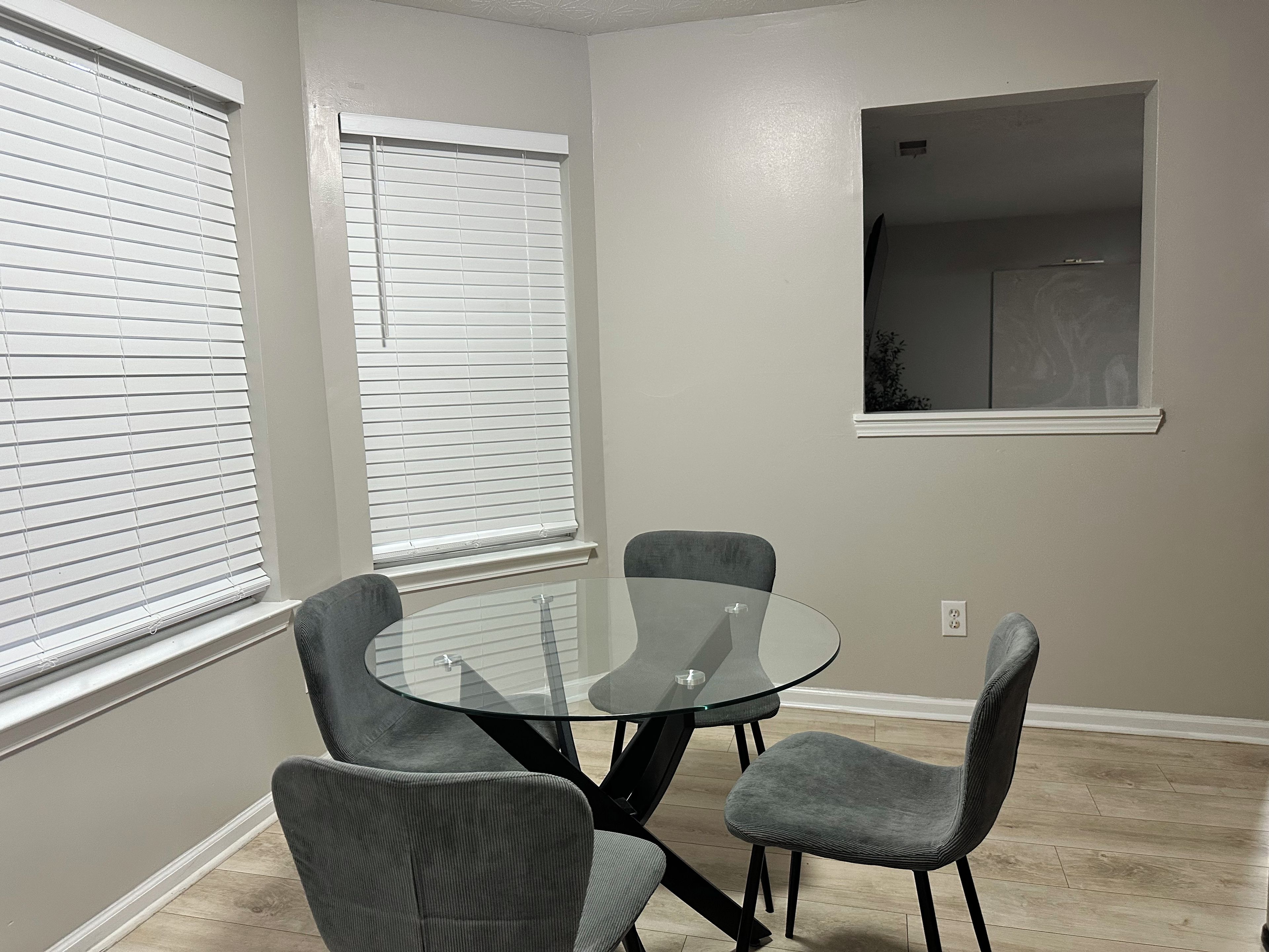 A dining area with a glass table and four grey chairs, two windows with blinds, and a recessed wall-mounted shelf.