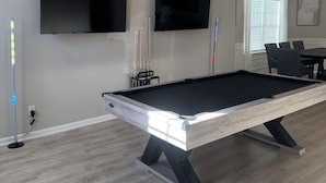 Ein Billardtisch mit modernem Design, ein an der Wand befestigter Flachbildfernseher und ein Essbereich mit Stühlen.