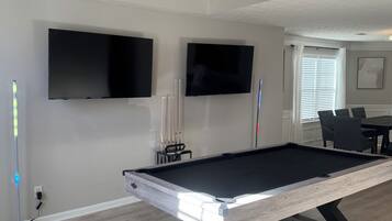 A pool table with a modern design, a flat-screen TV mounted on the wall, and a dining area with chairs.