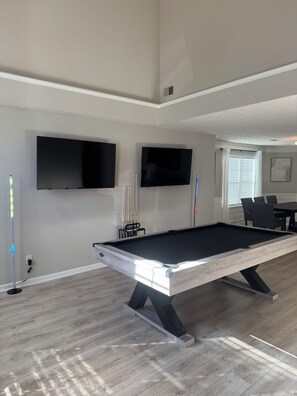 Ein Billardtisch mit modernem Design, ein an der Wand befestigter Flachbildfernseher und ein Essbereich mit Stühlen.