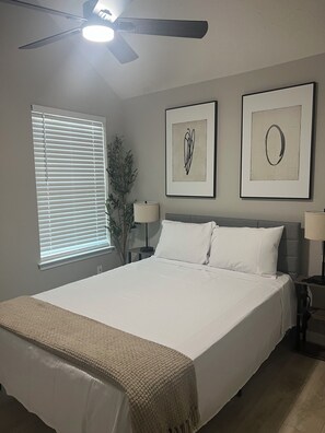 Ein Schlafzimmer mit einem Bett, einem Deckenventilator, einem Fenster mit Jalousien und zwei gerahmten Kunstwerken an der Wand.