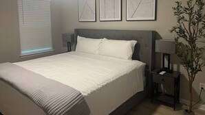 Ein Schlafzimmer mit einem Bett, einem Deckenventilator, einem Fenster mit Jalousien, einem Nachttisch mit Lampe und einer Wandpflanze.