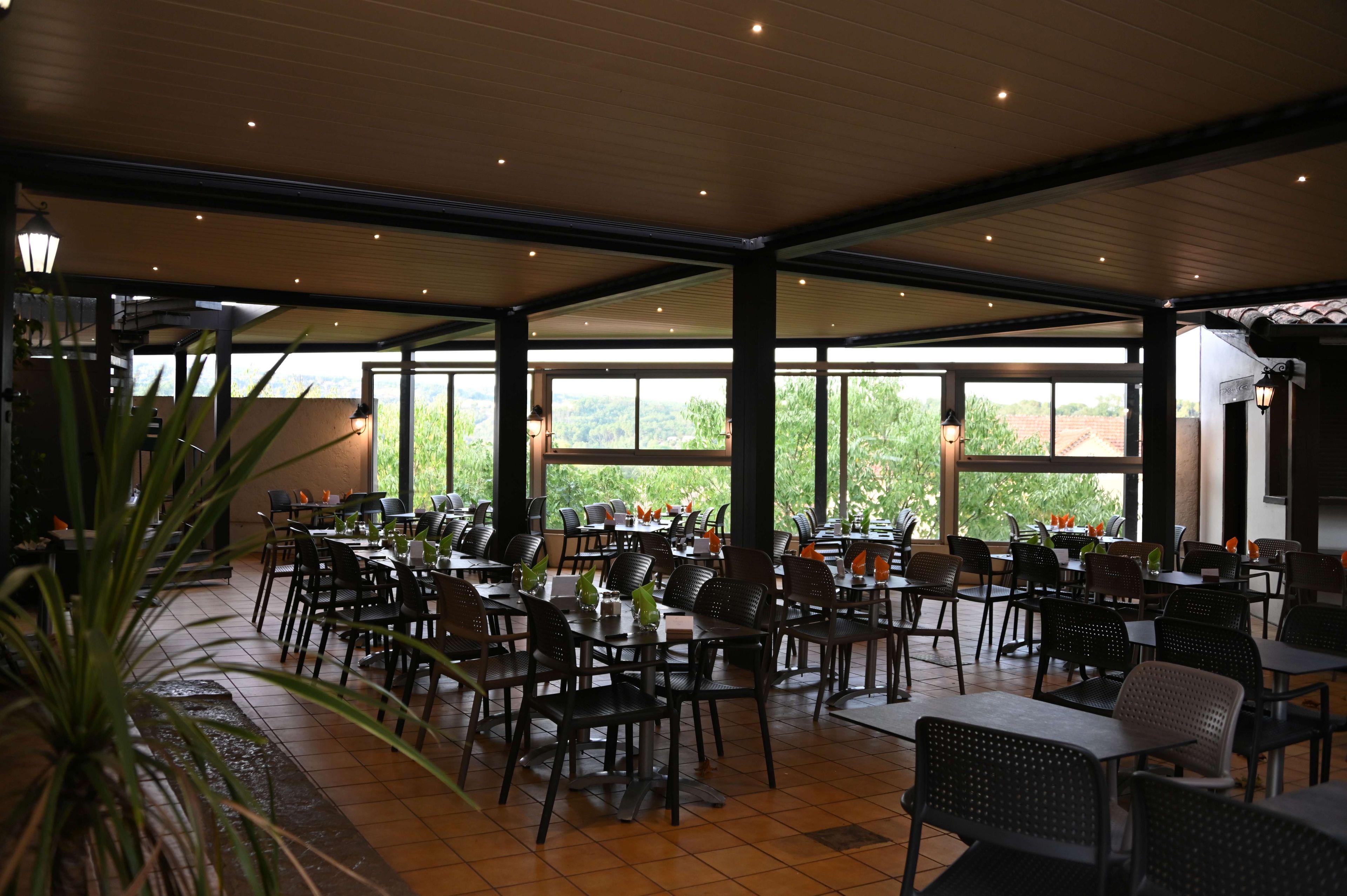 A spacious indoor dining area with multiple tables and chairs, large windows offering a view of greenery outside.