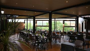 A spacious indoor dining area with multiple tables and chairs, large windows offering a view of greenery outside.