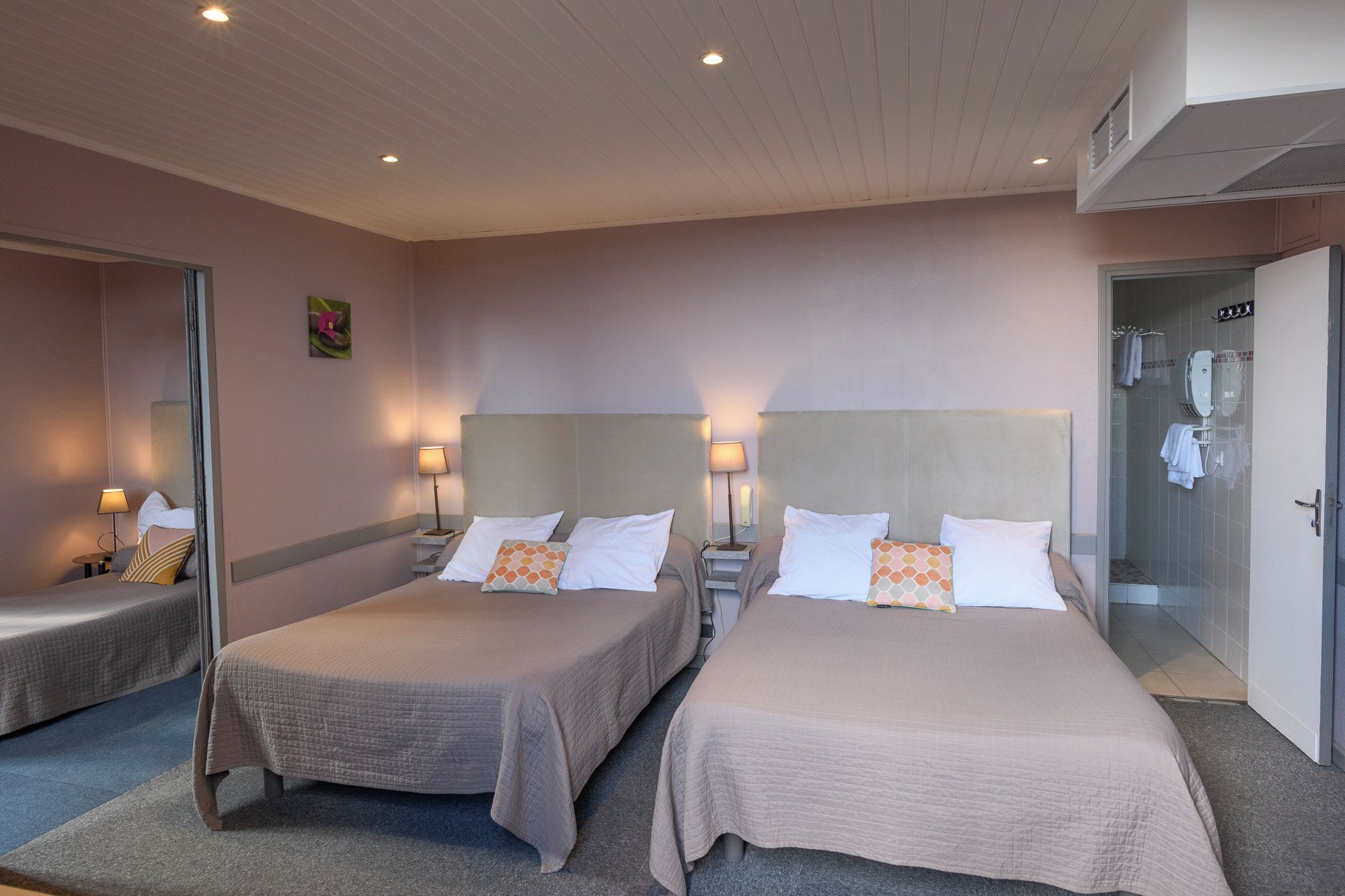 Quarto de hotel com duas camas, luminárias de cabeceira, área de chuveiro e espelho.