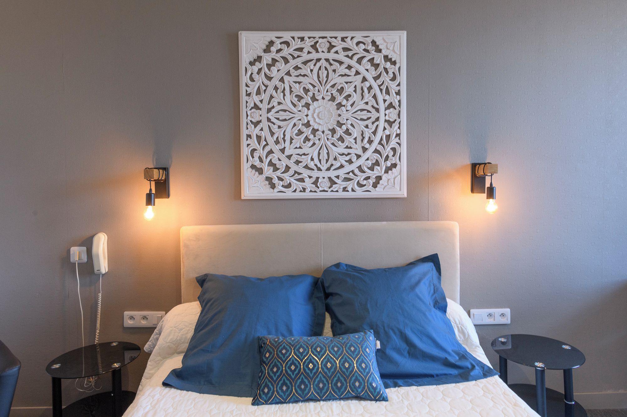 Une chambre d’hôtel comprenant un lit, deux tables de chevet, un luminaire fixé au mur et un élément décoratif mural.