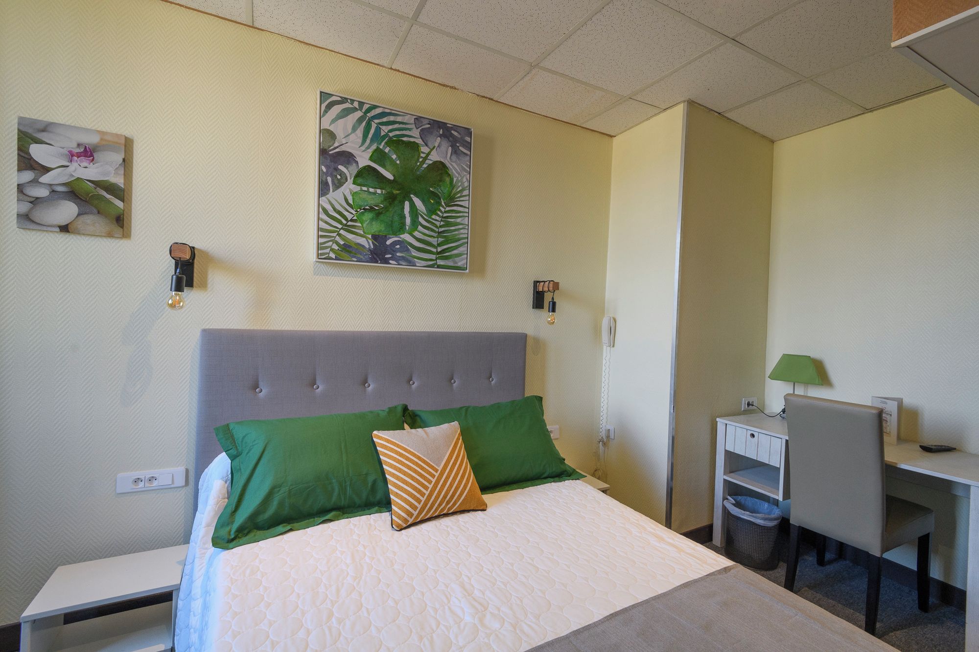 Habitación de hotel con cama, escritorio y silla. Hay dos lámparas fijadas a la pared y un teléfono.