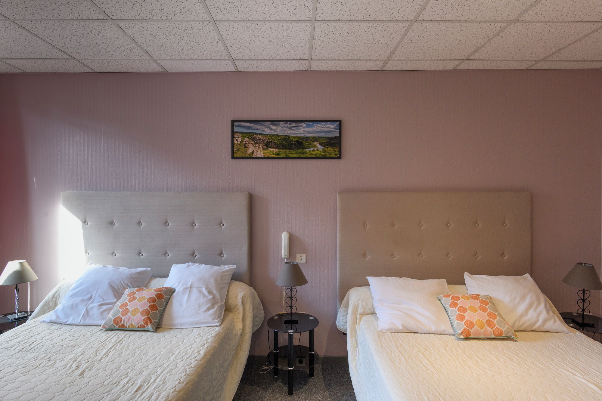 A hotel room with two beds, each with a headboard, a bedside table, a lamp, and a wall-mounted picture.