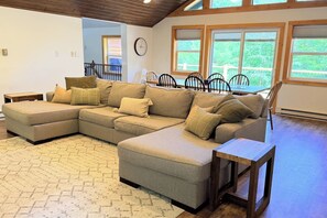 A sectional sofa with multiple cushions, a wooden coffee table, and a patterned rug in a room with large windows and a clock on the wall. - Last-Minute Fall Escape · Game Room & Fire Pit (Jay)
