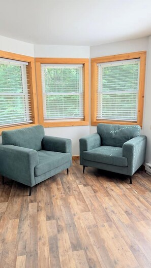 A room with two green armchairs, wooden flooring, and three windows with blinds. - Last-Minute Fall Escape · Game Room & Fire Pit (Jay)