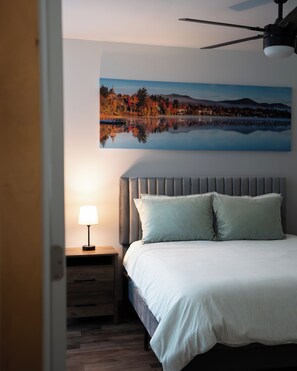 A bedroom with a bed, a nightstand, a lamp, a ceiling fan, and a wall-mounted painting of a landscape. - Whiteface Getaway | Fire Pit, Game Room, Bunk Room (Jay)