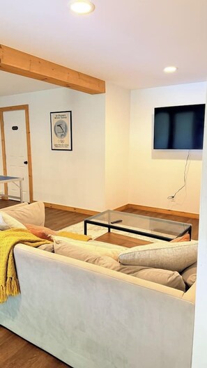 Een woonkamer met een bank, een glazen salontafel, een flatscreen televisie aan de wand en een houten balk in het plafond.
