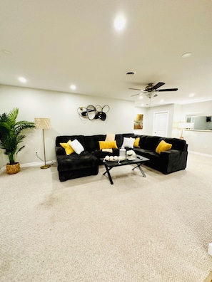 A modern living room with a black sofa, yellow and white cushions, a coffee table, a floor lamp, and a ceiling fan. - Cozy Retreat On The Eastside Estate (sleeps 4) (Greater Napanee)