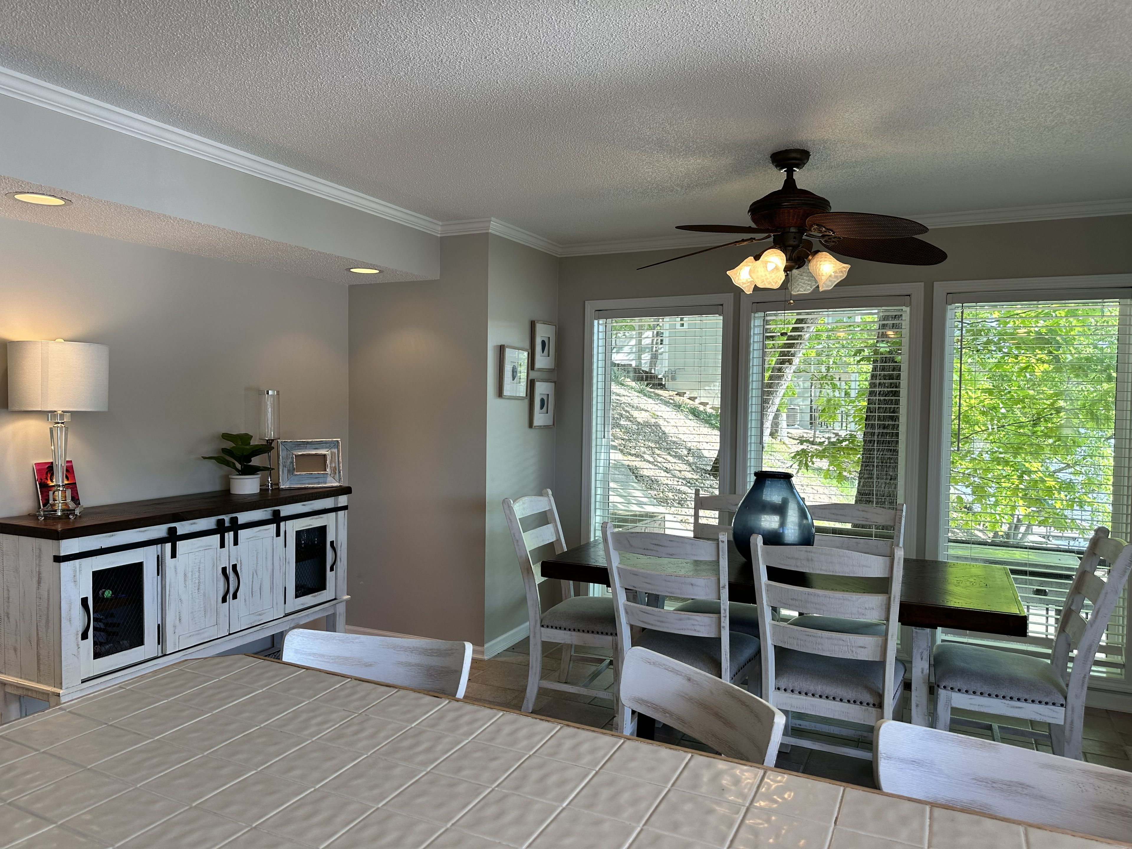 A dining area with a table, chairs, a ceiling fan, and a view of a green landscape through the windows.