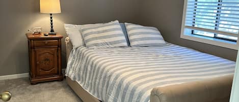 Une chambre à coucher comprenant un lit, une table de chevet, une lampe, un ventilateur de plafond et une fenêtre avec des stores.