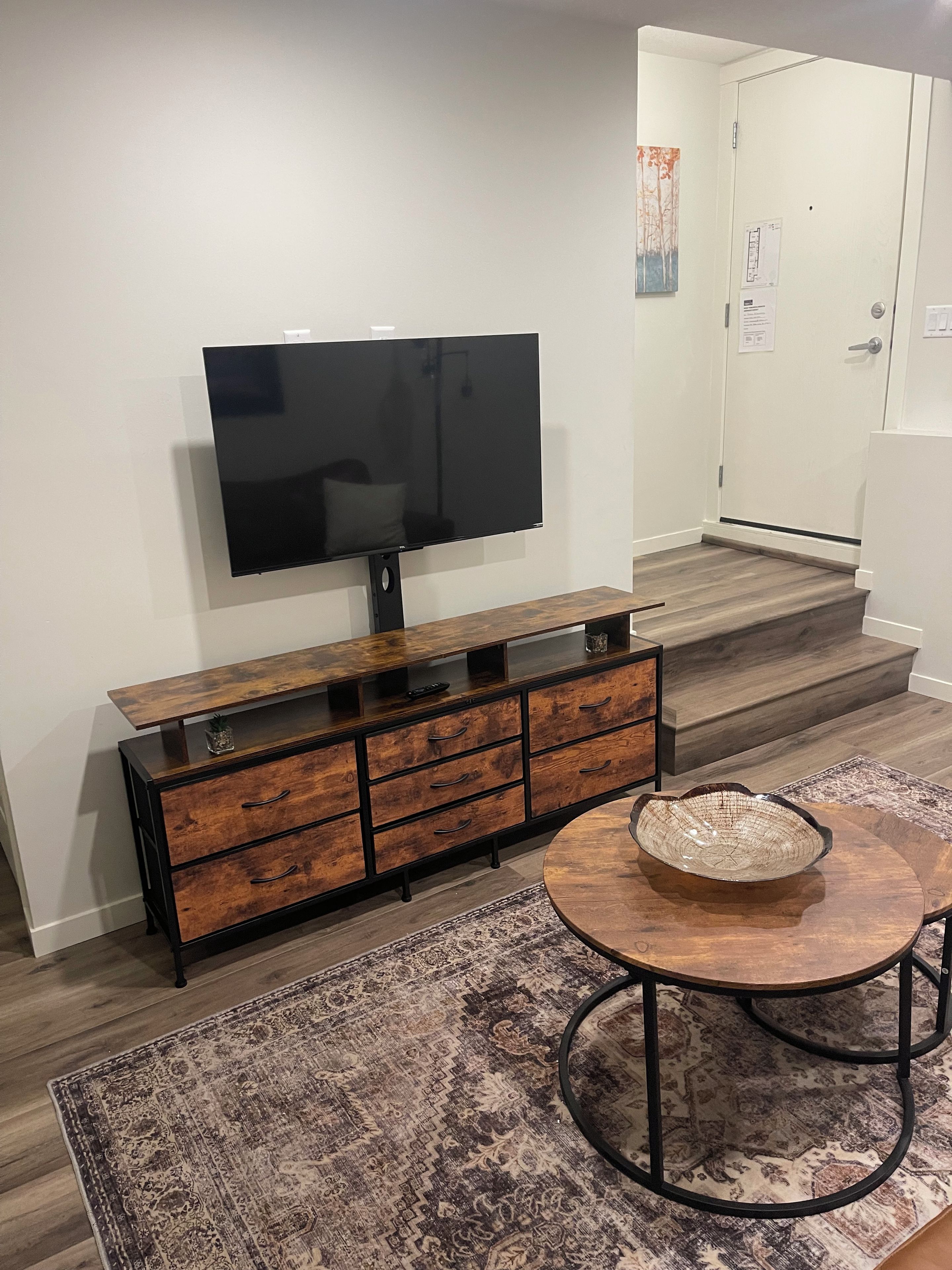 Un salon moderne avec un téléviseur à écran plat fixé à un meuble en bois, une table basse ronde surmontée d’un récipient et un tapis à motifs.